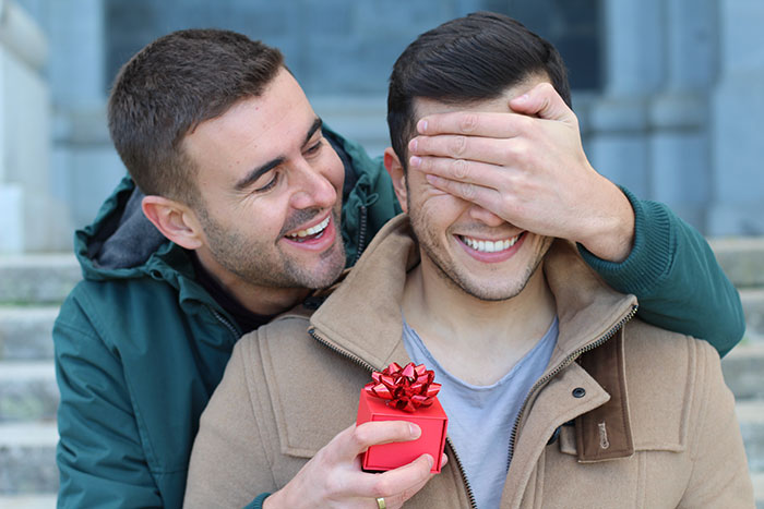 Couple gift giving, man behind other man holding hand over eyes with surprise gift